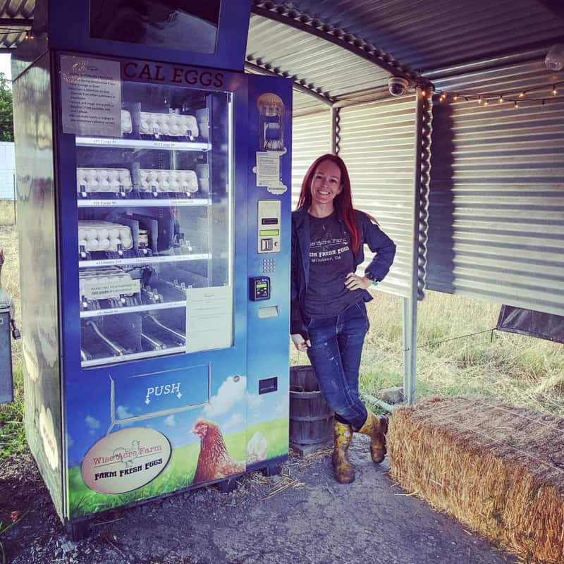 Vending machine selling eggs with a woman standing beside it in a farm setting.