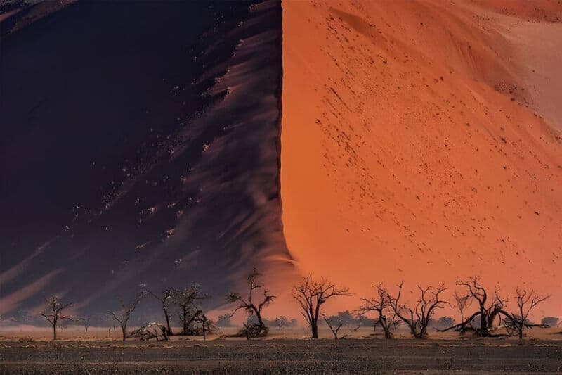 A massive sand dune with a sharp edge, trees in the foreground, and a dust storm.