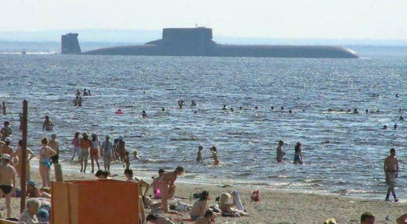 Beach scene with many people swimming and relaxing, large ship in the background.