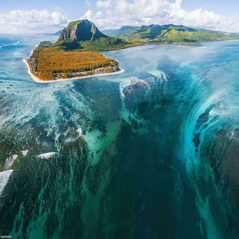 Aerial view of a large ocean wave crashing near a tropical island with mountains.