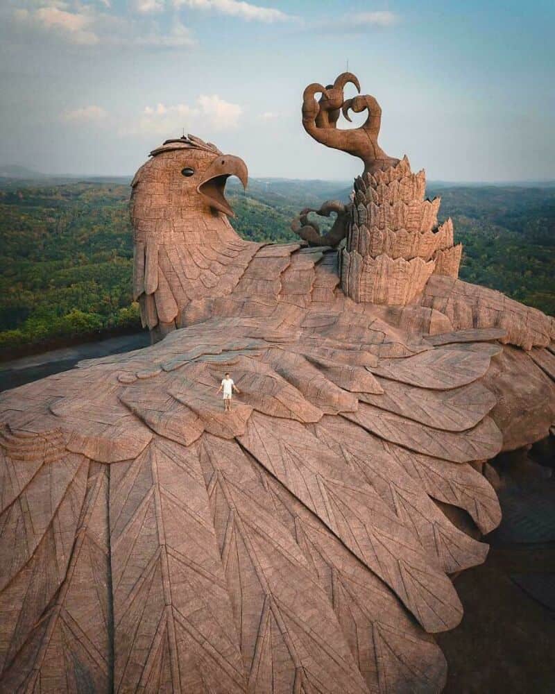 Large sculpture of a bird with a human figure standing on its wing, set in a landscape.
