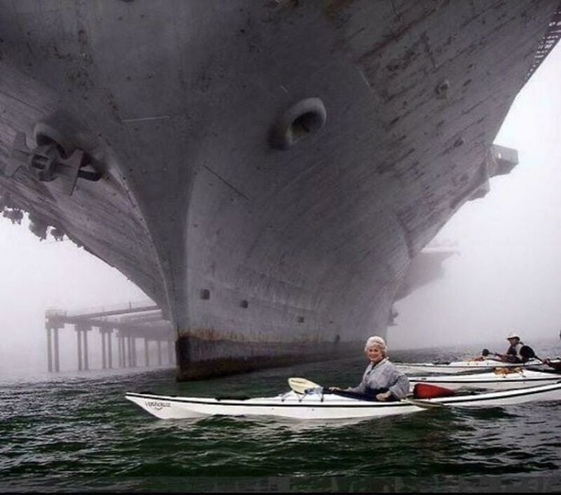 Large ship towering over kayaker in foggy water, with dock and other kayakers in background.