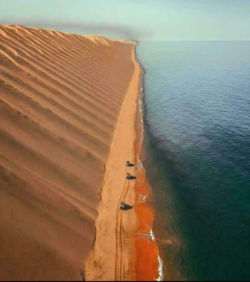 Aerial view of a large sand dune next to the ocean with vehicles on the sand.