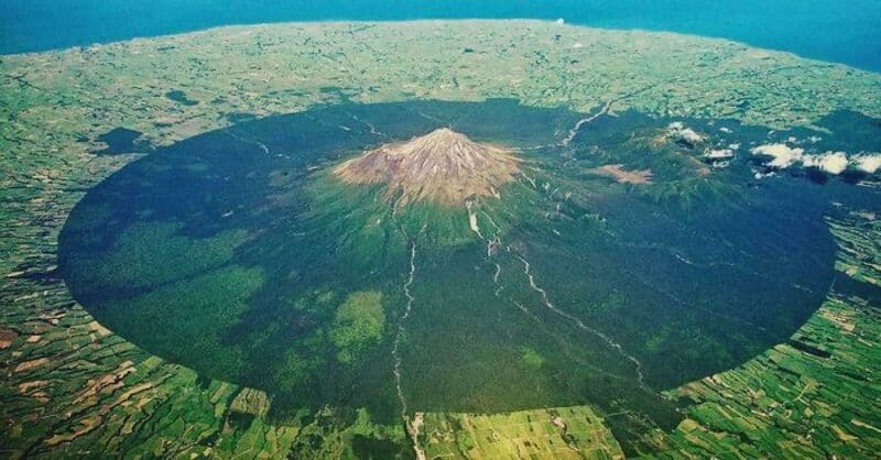 A large volcanic crater with a mountain in the center and surrounding green fields.