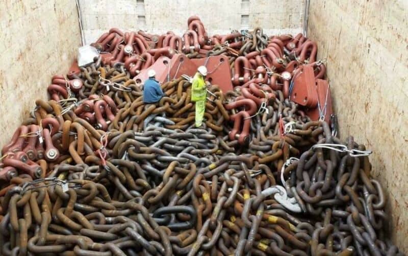 Two workers stand on a large pile of metal chains and pipes in a construction site.