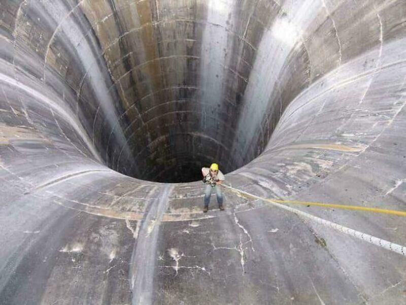 Person in yellow helmet and harness near large, deep, circular concrete structure.