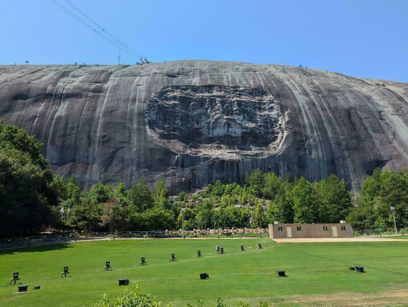 Large rock formation with a carved-out rectangular section, trees, and a grassy field.