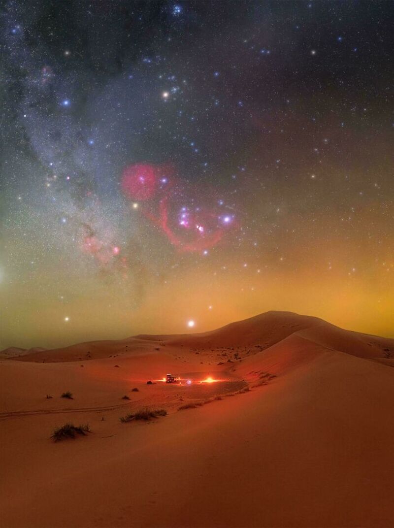 Starry night sky over desert with sand dunes and small campfire.