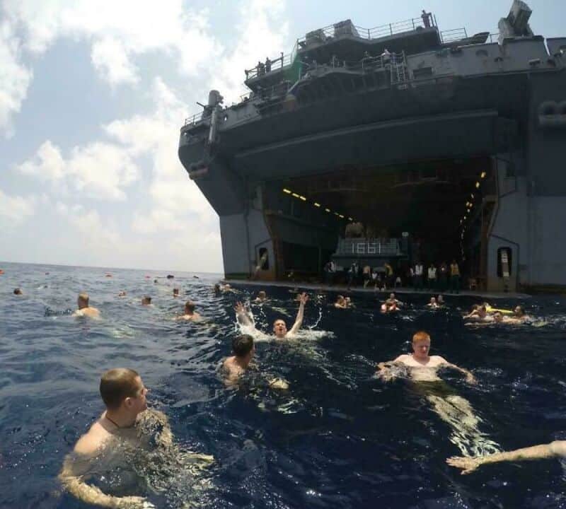 People swimming near a large aircraft carrier at sea.