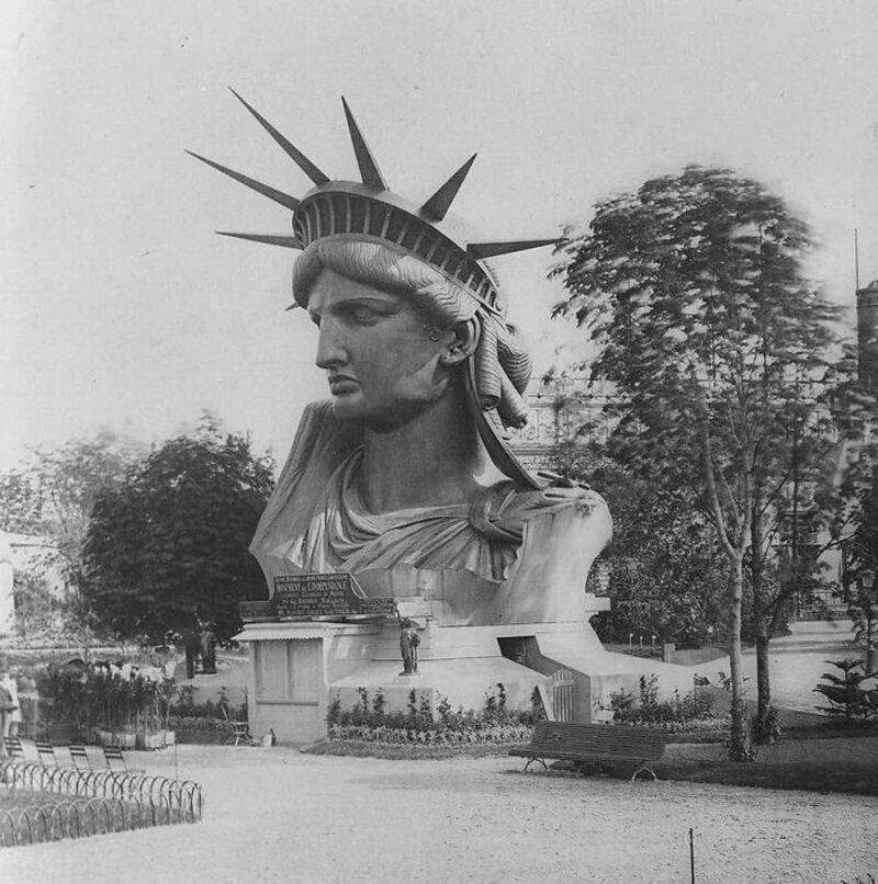 Black and white photo of a giant Statue of Liberty head in a park.