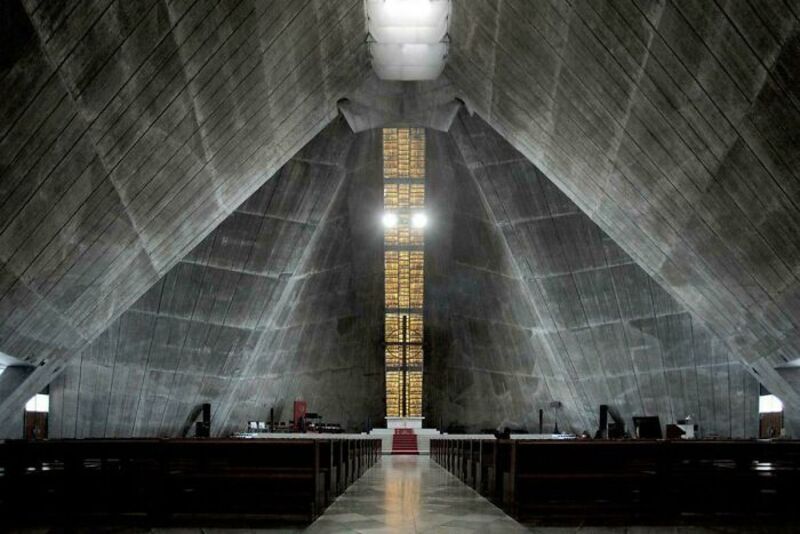 Interior of a large, concrete church with a high, arched ceiling and a tall, narrow window.