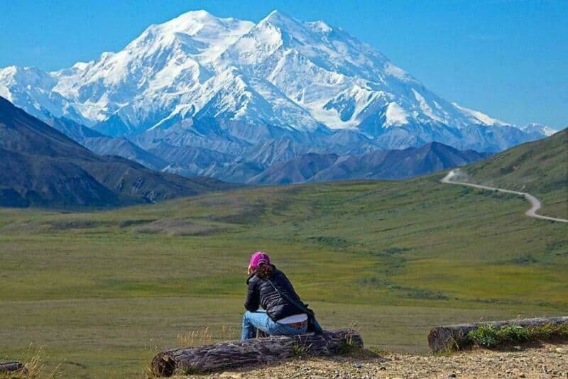 Person sitting on a log looking at snow-capped mountains and green hills.