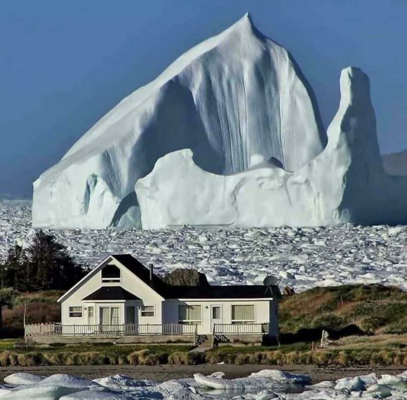 Large iceberg towering over a house in a snowy landscape.