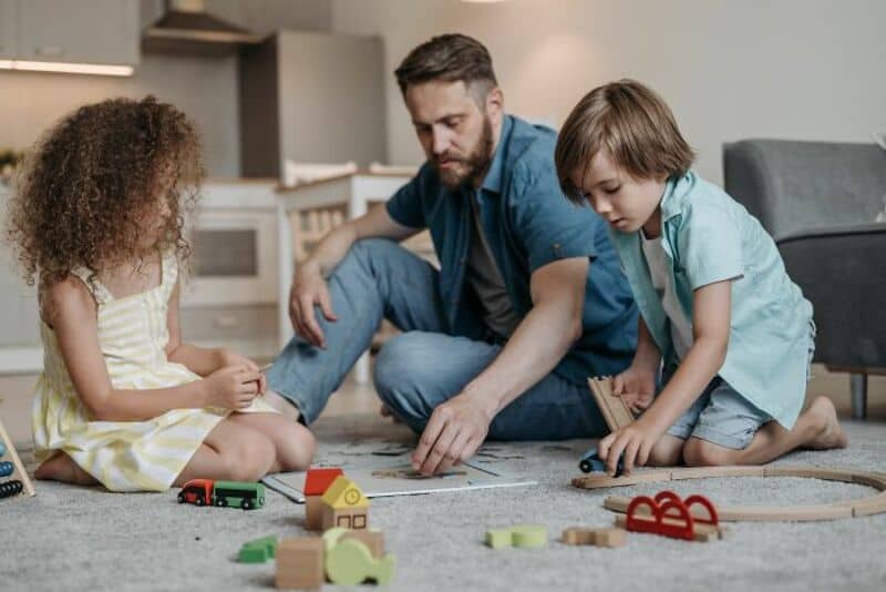 Father playing with two children on the floor with toys in a living room.