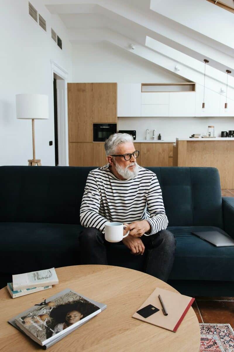 Older man with glasses and a striped shirt sitting on a dark sofa holding a mug.