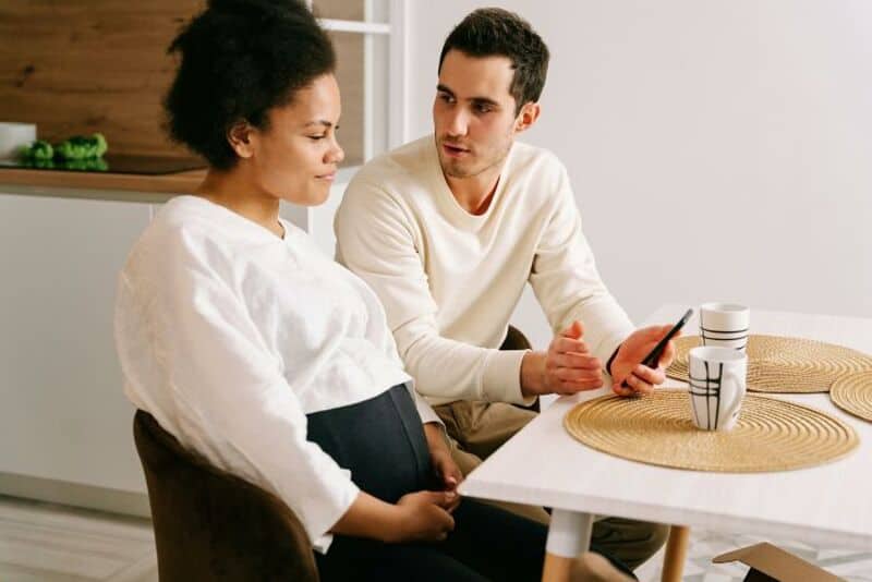 A man and pregnant woman sitting at a table in a kitchen, talking.