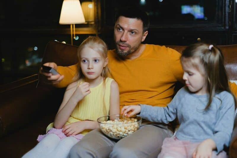 Father watching TV with two young daughters, one eating popcorn, at home.