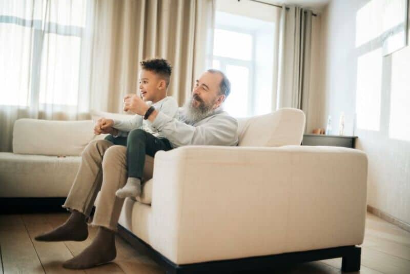 Father and son playing and laughing on a sofa in a bright living room.