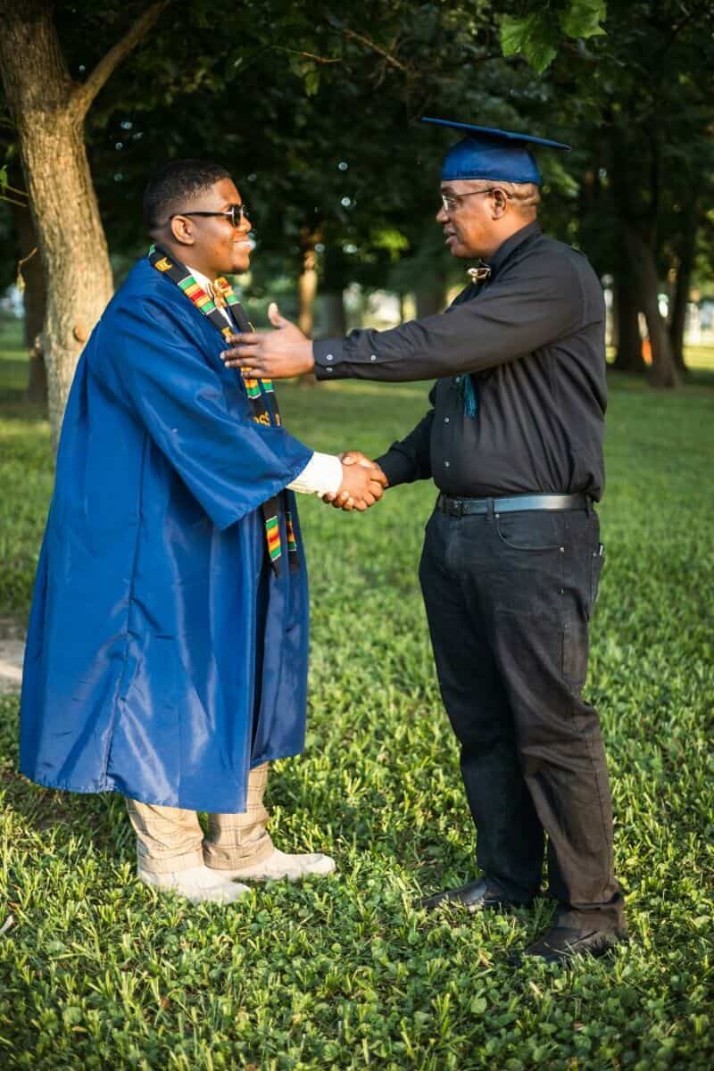 Graduate in cap and gown shaking hands with man in black shirt outdoors.