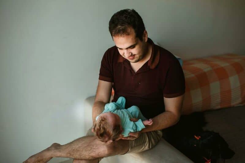 Father holding baby in a dimly lit room, with a bed in the background.