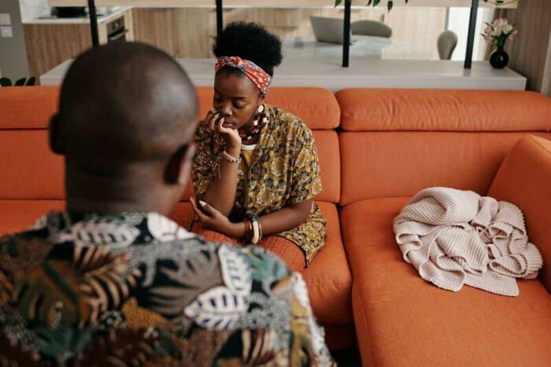 A girl with a headband and patterned dress sits on a sofa, talking to a man with a shaved head.