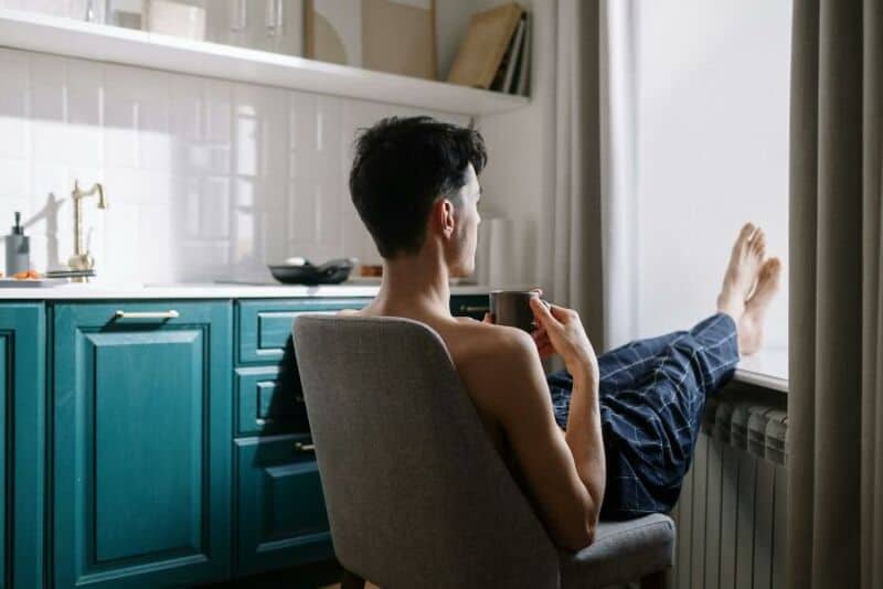 Young man relaxing with coffee at home, sitting by window with blue kitchen cabinets.