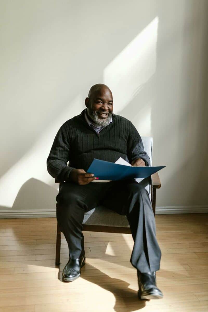 Smiling man sitting on a chair with a document in hand, sunlight on wall.