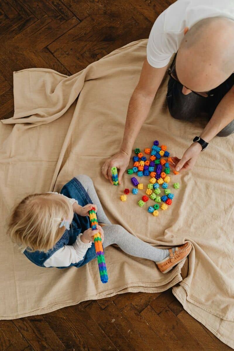 A man and a child playing with colorful building blocks on a beige blanket.