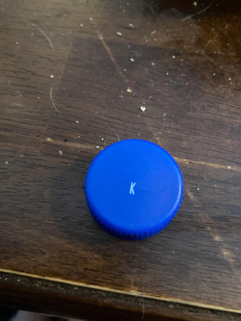 Blue bottle cap with a white letter K on a wooden surface.
