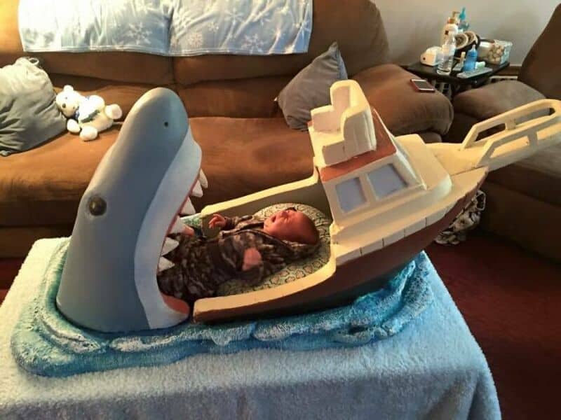 Baby in a boat with a shark and whale plush toys in a living room.
