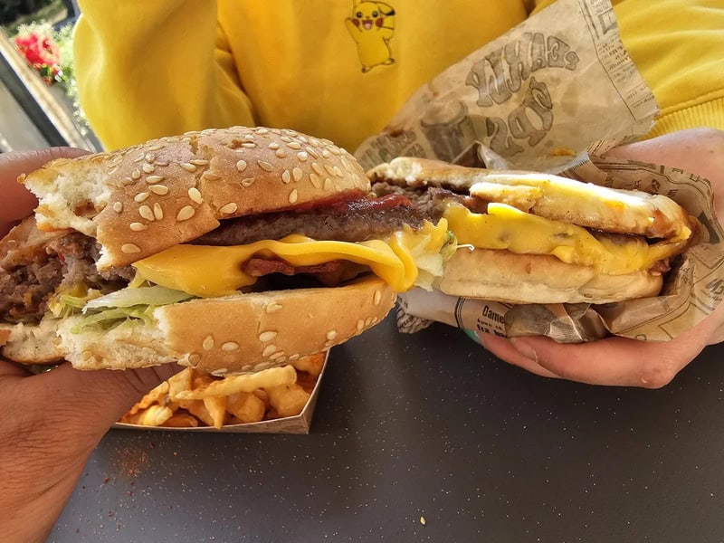 Two hands holding a cheeseburger and a breakfast sandwich in a fast food restaurant.
