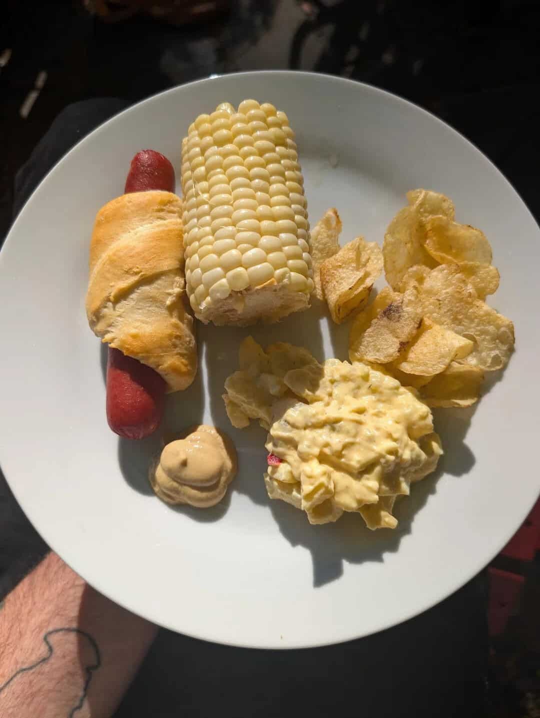 Plate with corn on the cob, potato chips, a hot dog with mustard, and a dollop of mustard.