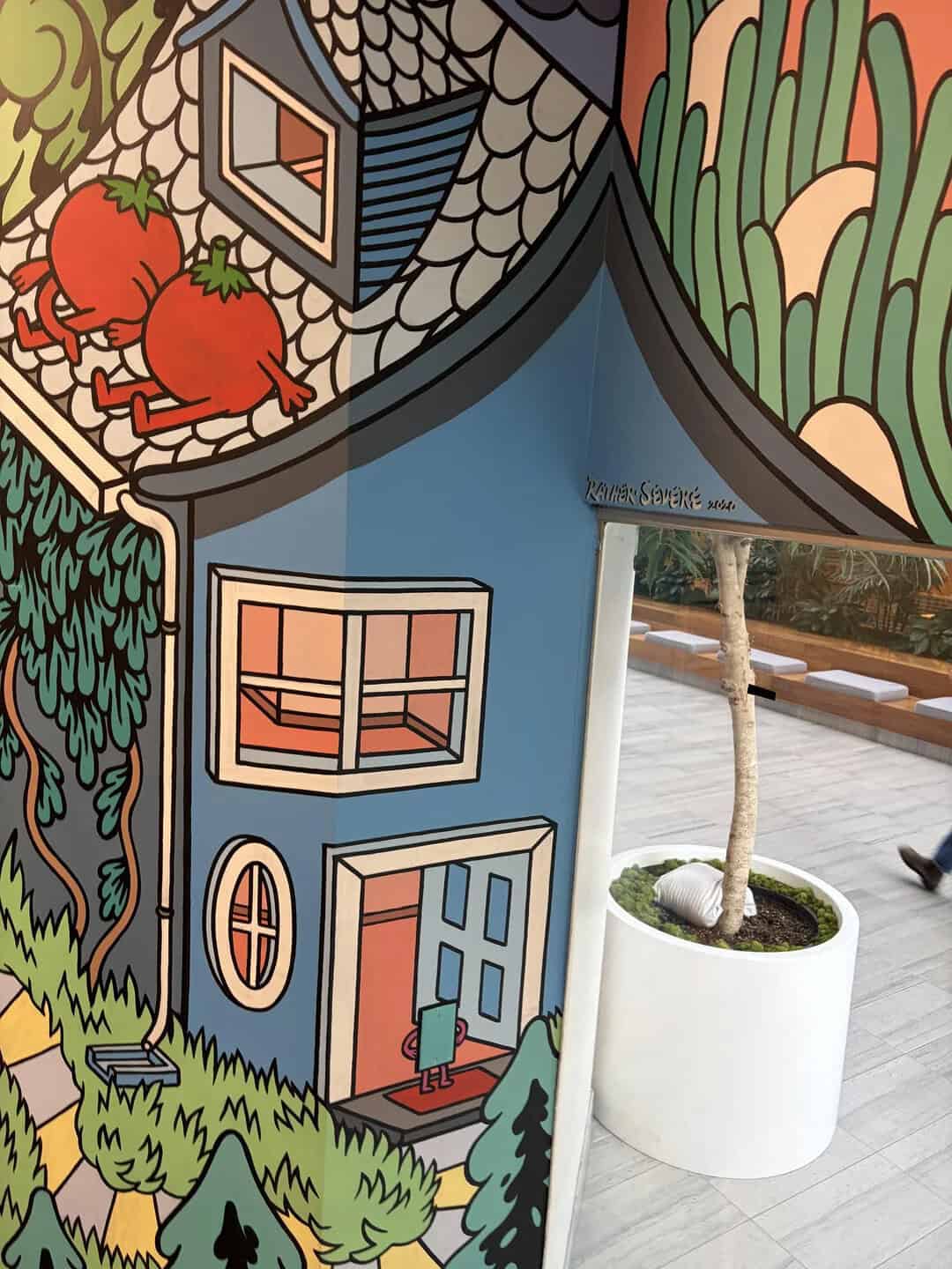Colorful mural of a house with a window, door, and trees, next to a real potted tree.