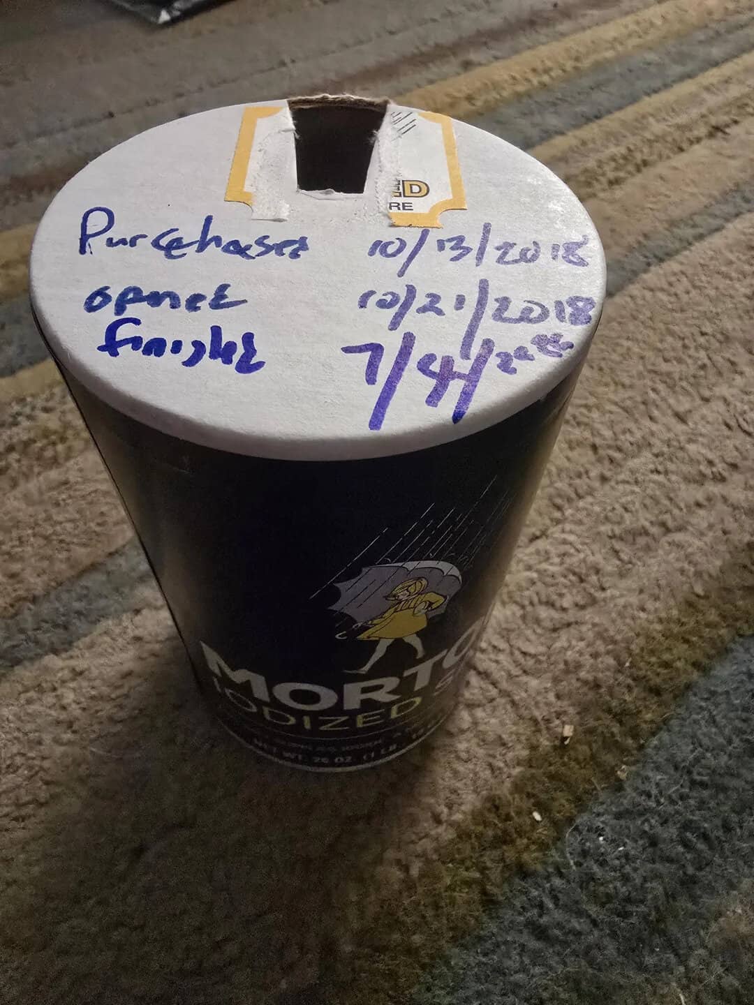 A black cylindrical object with a white top, handwritten notes, and a yellow sticker, on a textured,.