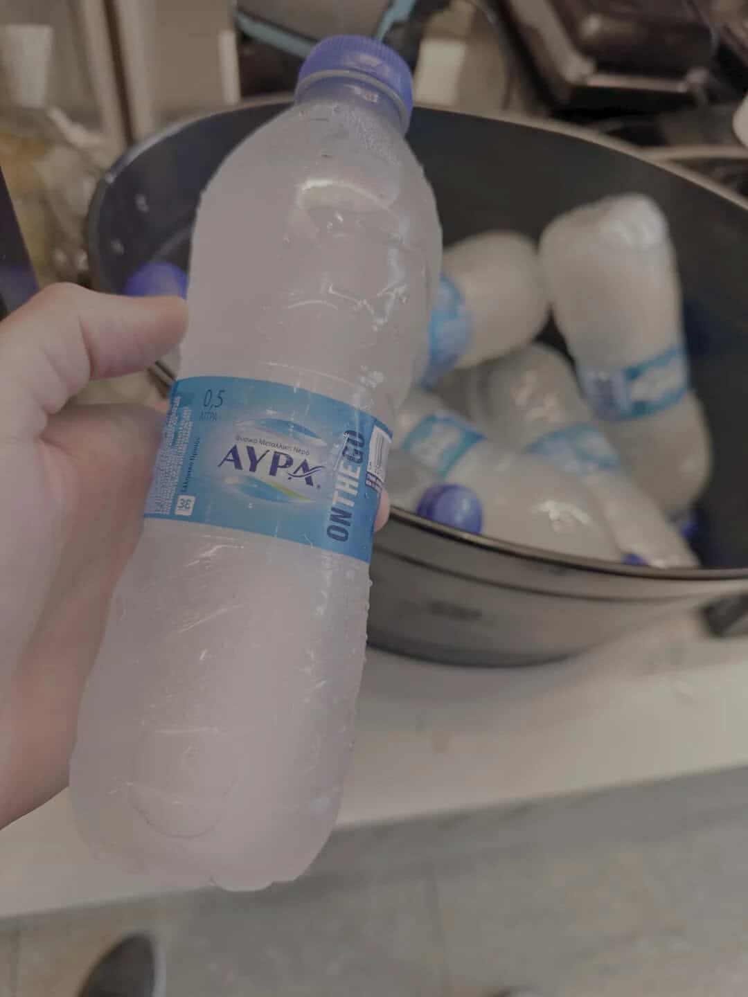 Hand holding a small water bottle with a blue cap, with a bucket of similar bottles in the back.