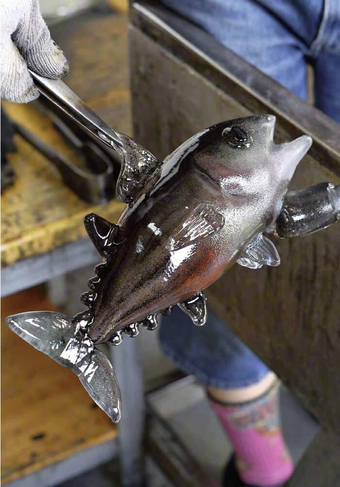 Glass fish sculpture being held with pliers, in a workshop setting.