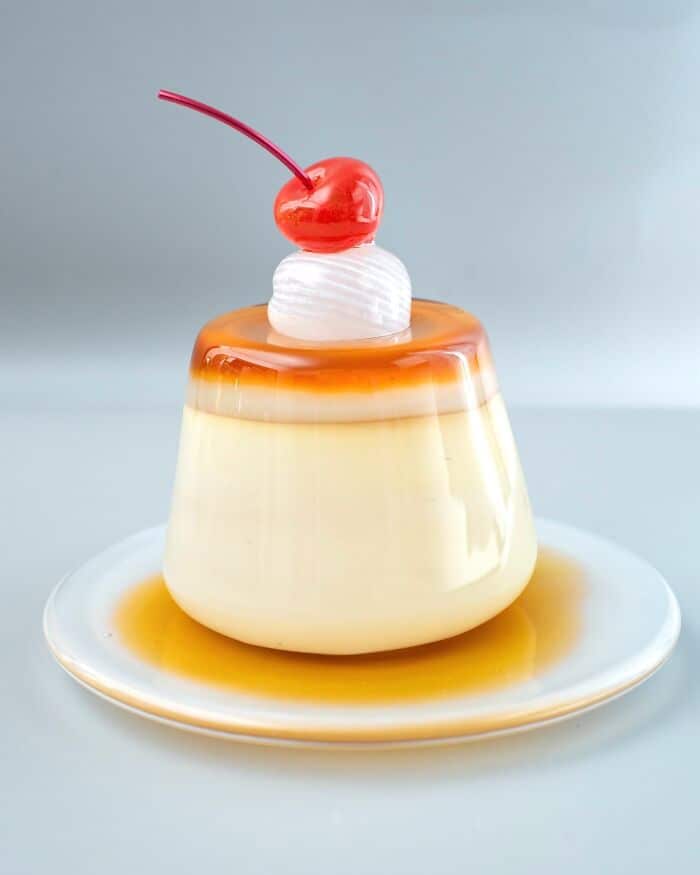 Creamy caramel flan with cherry and whipped cream on a white plate.