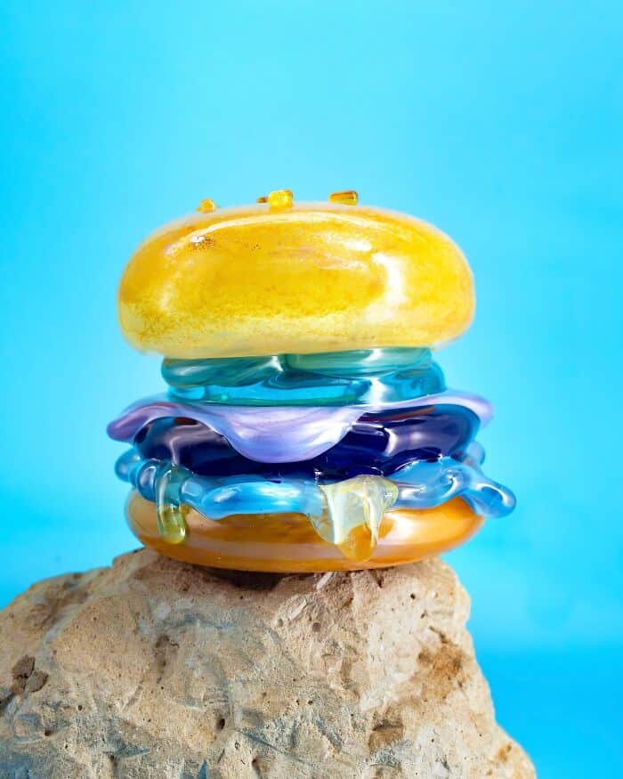 Colorful glass food sculptures stacked on a rock against a blue background.
