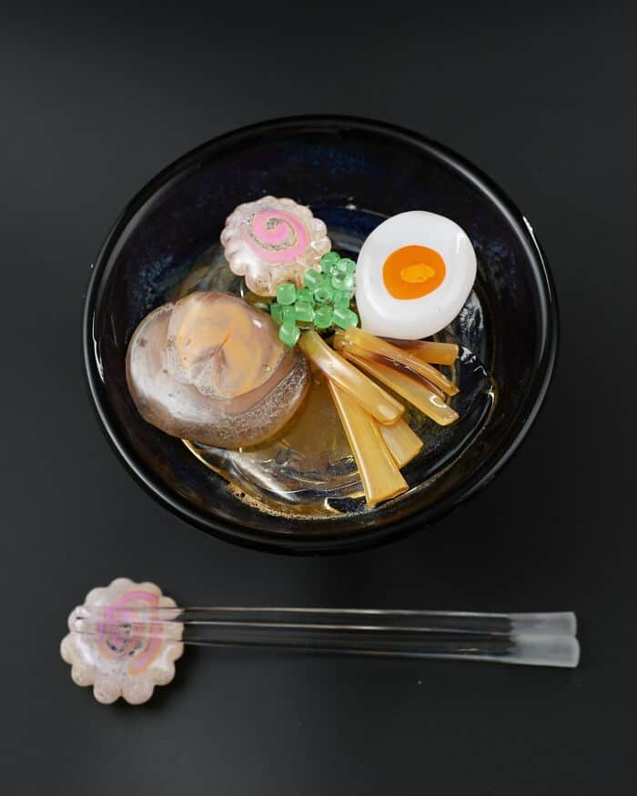Glass food sculptures including a fried egg, ramen, and a pink swirl dessert in a black bowl.