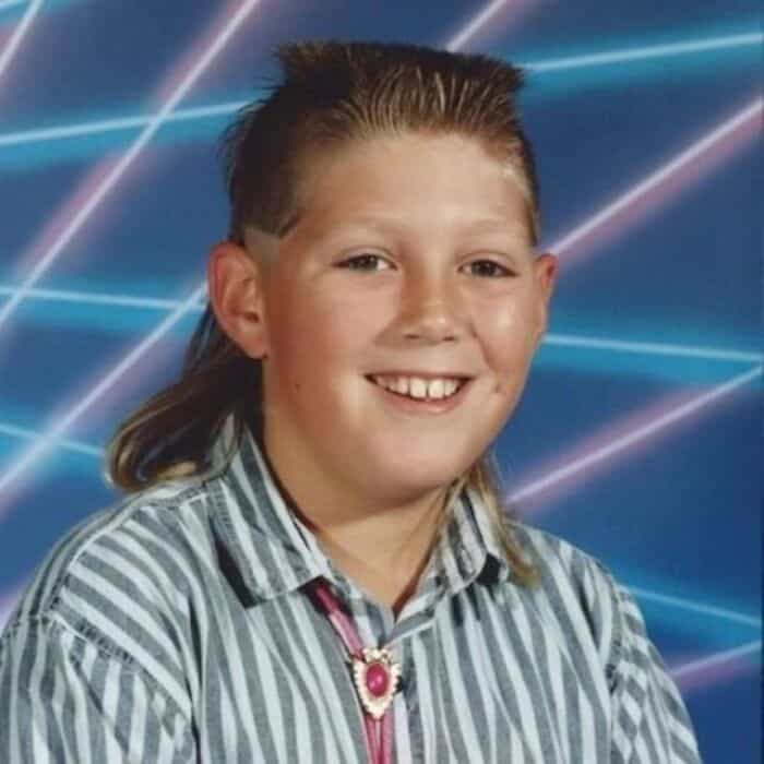 Child with mullet hairstyle, smiling, wearing a striped shirt and a pink necklace.