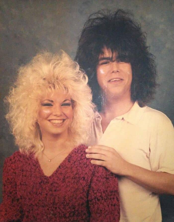Two people with big, curly 80s hairstyles, smiling, one in a red sweater, the other in a white shirt.