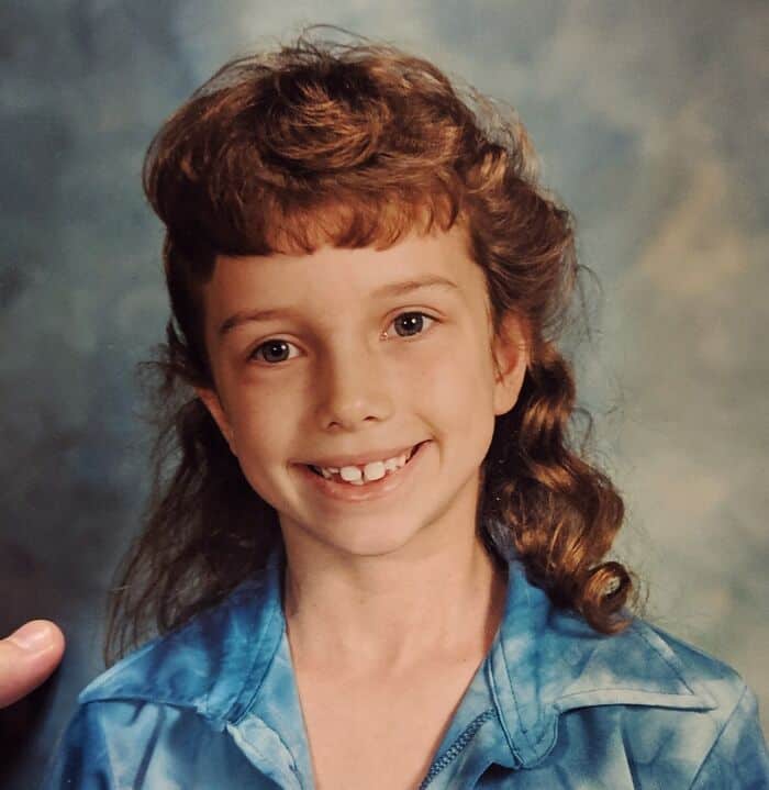 Child with curly mullet hairstyle, smiling, wearing a blue shirt.