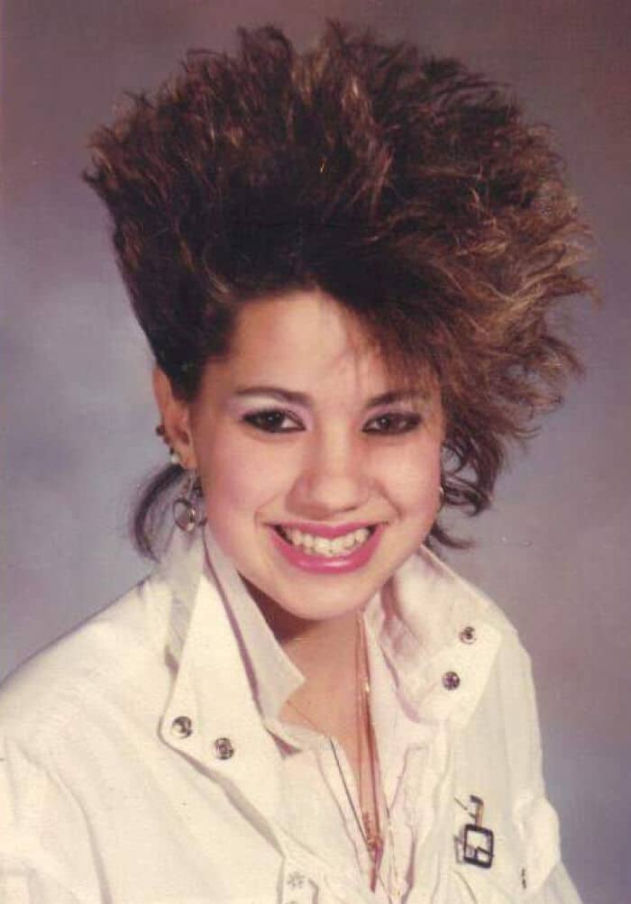 Woman with big, teased 80s/90s hairstyle, smiling, wearing a white jacket.