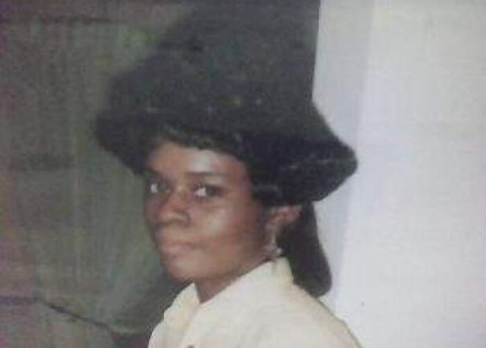 Woman with a large, curly black hat and a vintage hairstyle from the 80s or 90s.