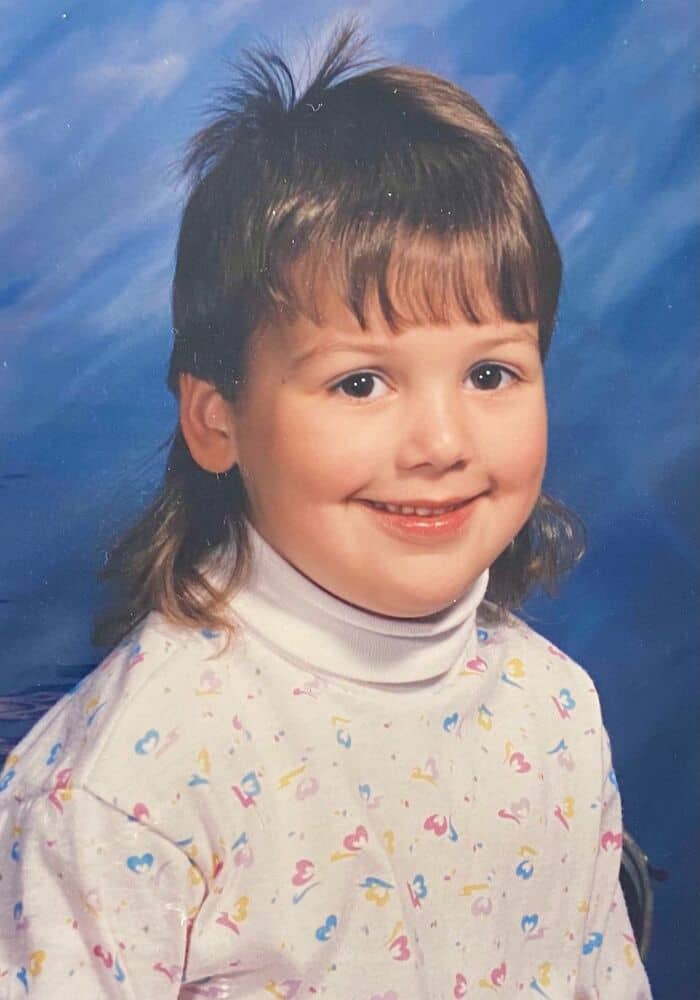 Child with mullet hairstyle, smiling, wearing a colorful patterned shirt.