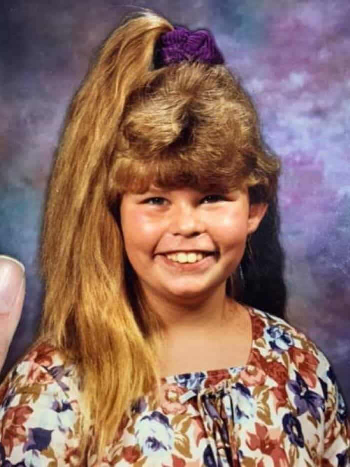 Girl with big 80s/90s hairstyle, long blonde hair with a purple scrunchie, smiling.
