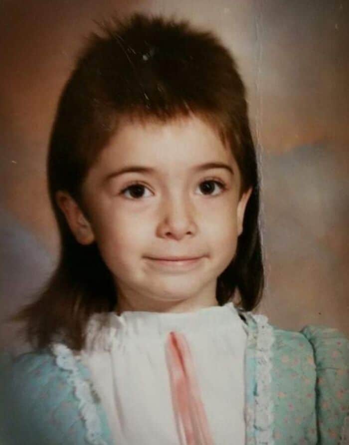 Child with 80s/90s hairstyle, mullet, smiling, wearing a white and light blue dress.