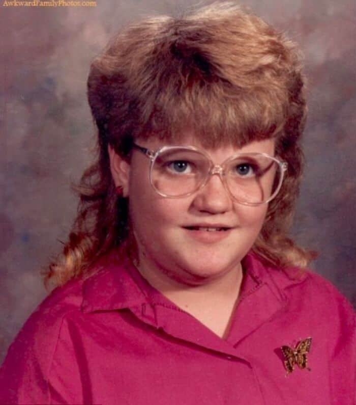 Child with big 80s/90s hairstyle, glasses, pink shirt, butterfly pin.