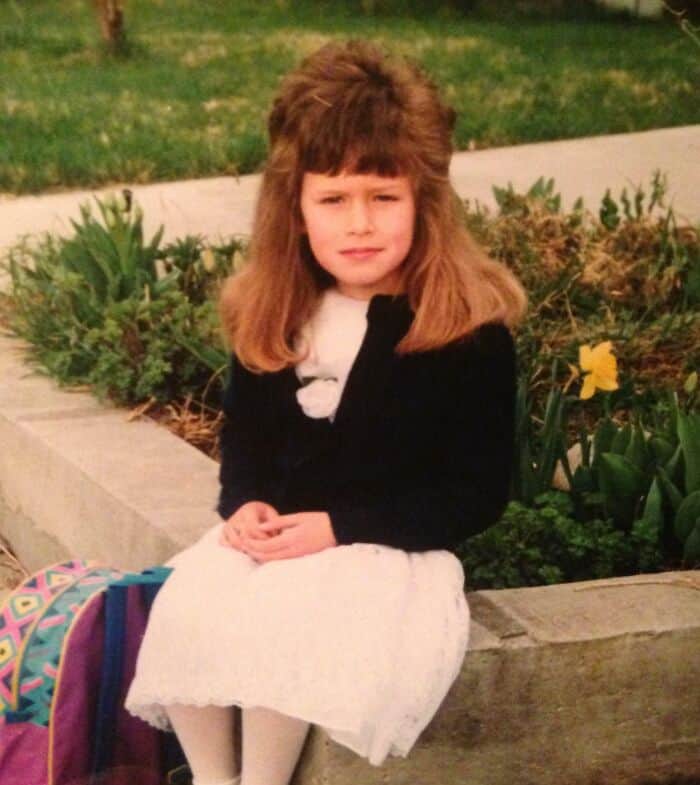 Young girl with big, voluminous 80s-style hair, sitting outdoors with a garden background.