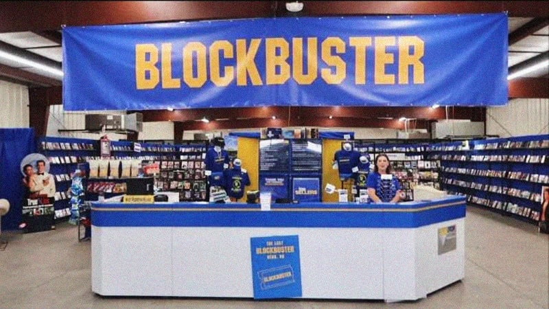 Inside a store with a large 'BLOCKBUSTER' banner, shelves filled with DVDs, and staff at the counter.